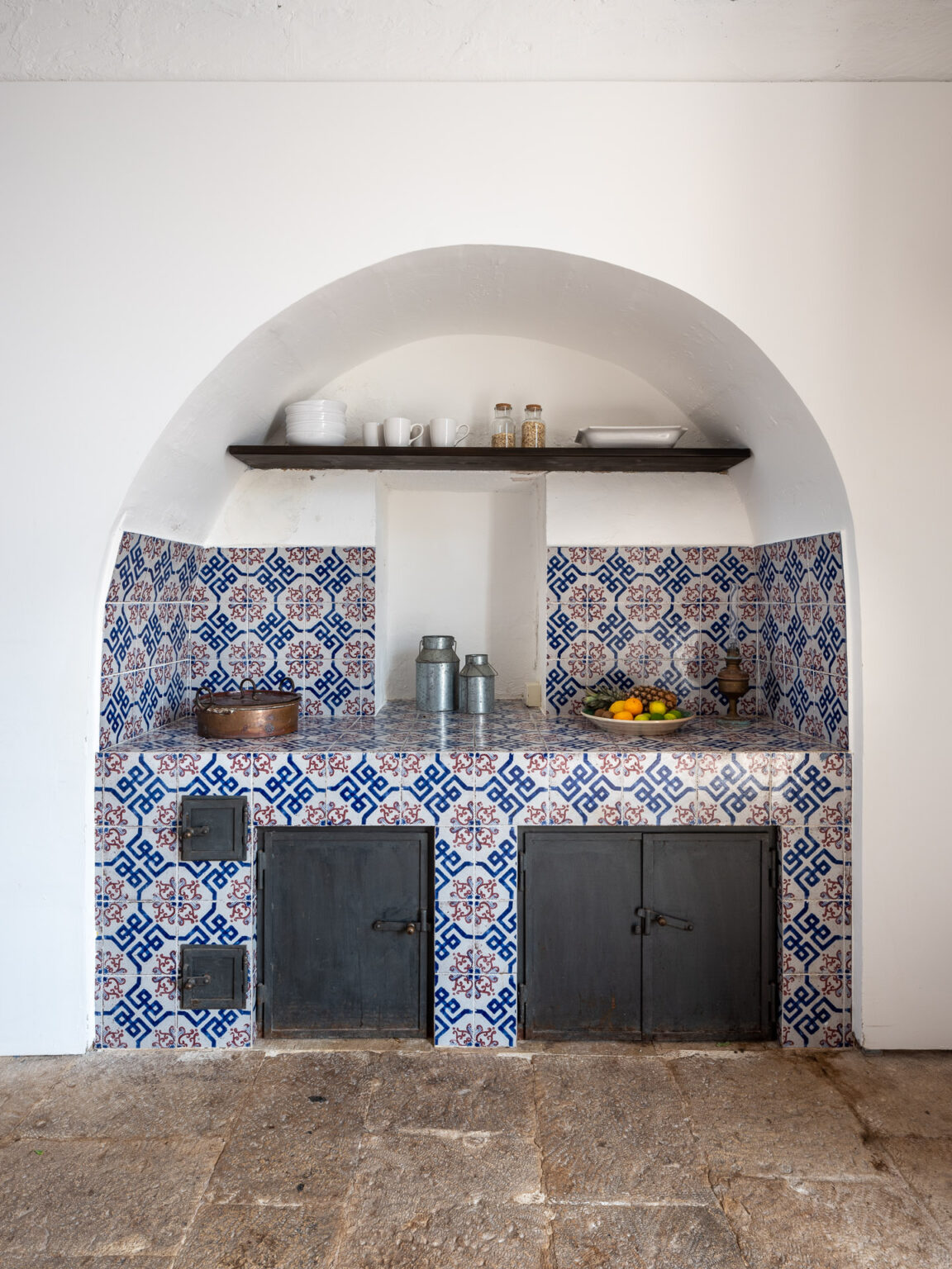 Charming kitchen nook featuring blue and white patterned tiles, a wooden shelf with dishes and jars, and a countertop displaying fresh fruits. The design includes two black cabinets below, set against a light-colored wall, creating a warm and inviting atmosphere. | Only on SopranoVillas