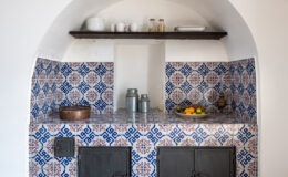 Charming kitchen nook featuring blue and white patterned tiles, a wooden shelf with dishes and jars, and a countertop displaying fresh fruits. The design includes two black cabinets below, set against a light-colored wall, creating a warm and inviting atmosphere.