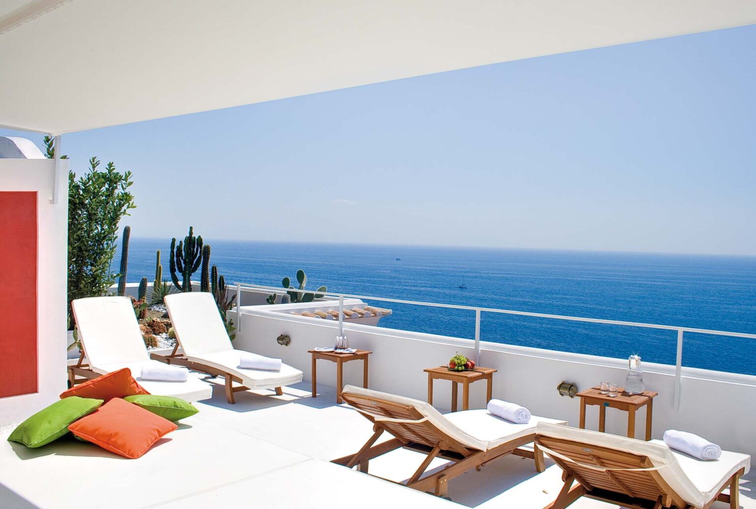 Luxurious rooftop terrace with sun loungers, colorful cushions, and wooden tables overlooking the serene blue sea, featuring tropical plants and a clear sky, ideal for relaxation and leisure. | Only on SopranoVillas
