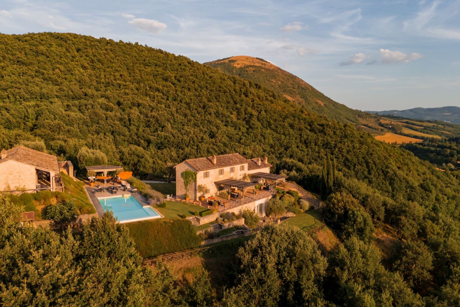 Aerial view of a rustic villa surrounded by lush green hills, featuring a swimming pool and outdoor seating areas, ideal for relaxation and vacation in a picturesque landscape. | Only on SopranoVillas