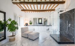 Spacious modern bathroom featuring a glass shower, marble flooring, and wooden beams, with a comfortable chaise lounge and stylish decor elements.