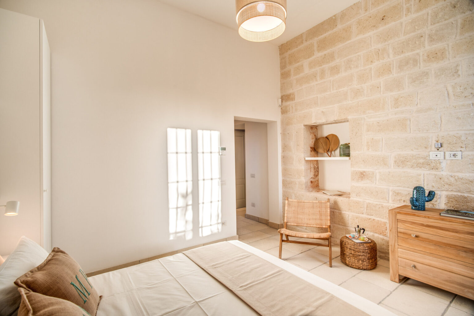 Bright and airy bedroom featuring natural stone walls, a wooden dresser, a cozy chair, and large windows that allow ample sunlight. The minimalist decor creates a serene atmosphere, ideal for relaxation. | Only on SopranoVillas