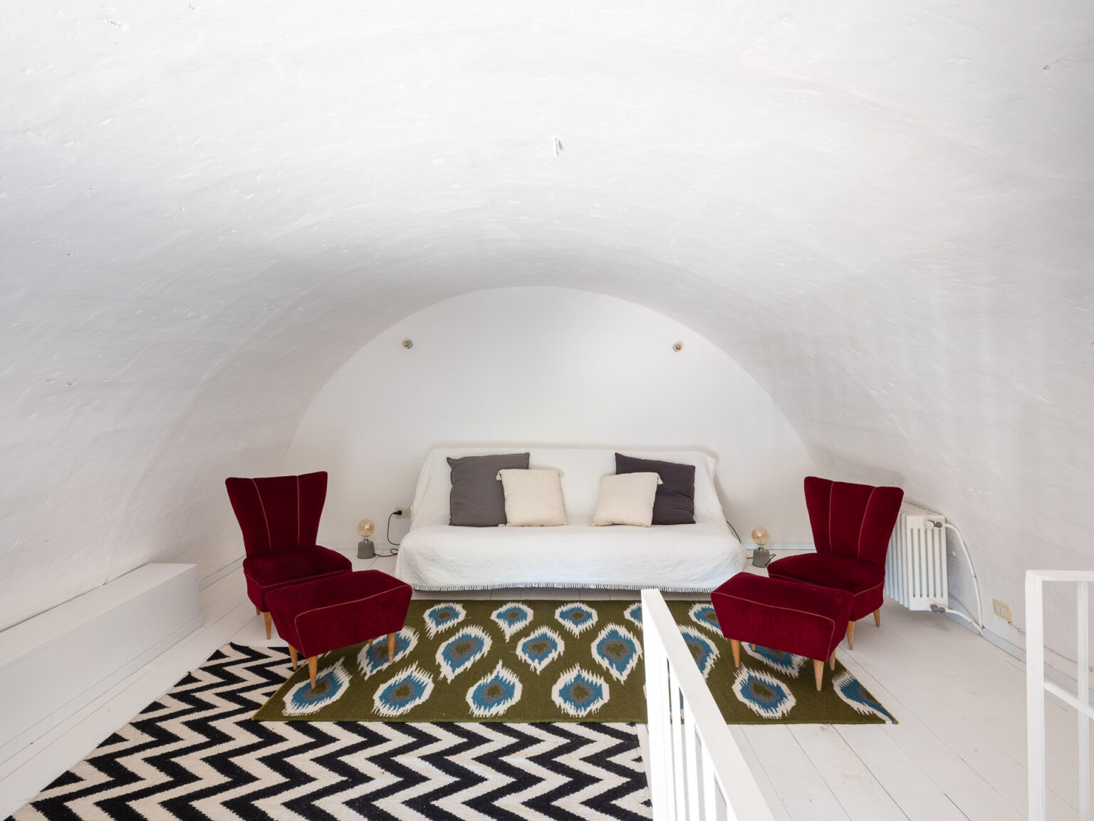 Cozy attic living space featuring a white curved ceiling, a minimalist white sofa with decorative cushions, and two red velvet chairs. The room is adorned with a patterned area rug and has a bright, airy feel. | Only on SopranoVillas