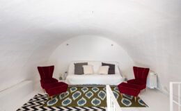 Cozy attic living space featuring a white curved ceiling, a minimalist white sofa with decorative cushions, and two red velvet chairs. The room is adorned with a patterned area rug and has a bright, airy feel.