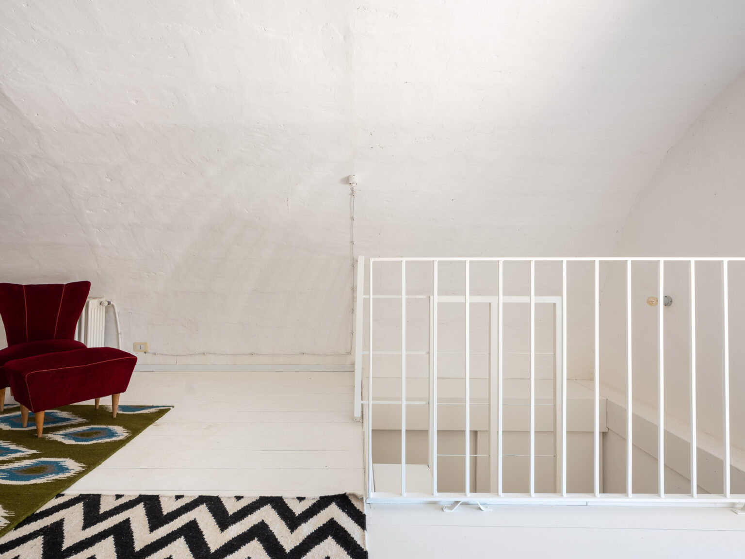 Cozy attic space featuring a red velvet armchair, geometric patterned rugs, and a white railing, highlighting a minimalist design and bright, airy atmosphere. | Only on SopranoVillas