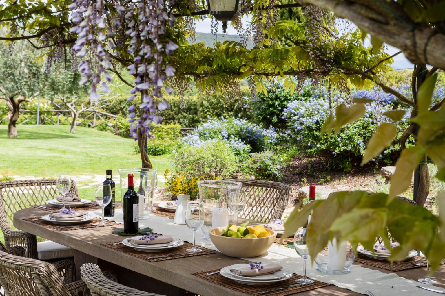 Outdoor dining setup featuring a rustic table adorned with plates, wine glasses, and a bowl of fresh lemons, surrounded by lush greenery and blooming flowers. Wisteria vines hang overhead, creating a serene and inviting atmosphere for al fresco meals. | Only on SopranoVillas