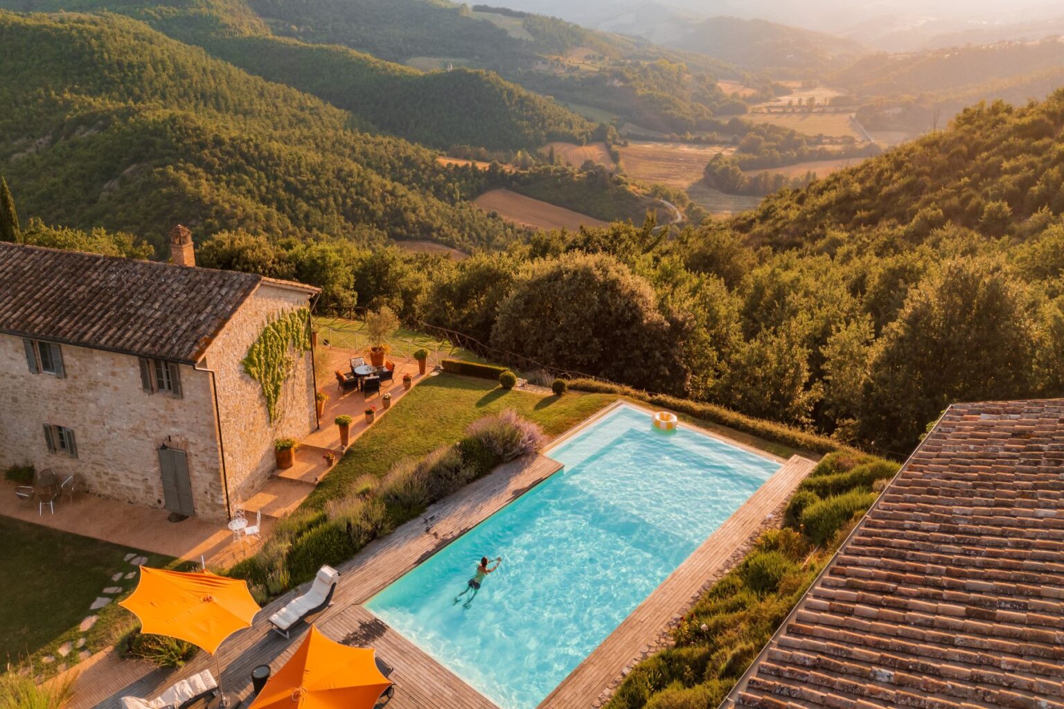 Aerial view of a charming stone villa with a swimming pool surrounded by lush green hills, featuring orange umbrellas and a relaxing outdoor seating area, ideal for a tranquil getaway in nature. | Only on SopranoVillas