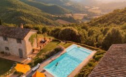 Aerial view of a charming stone villa with a swimming pool surrounded by lush green hills, featuring orange umbrellas and a relaxing outdoor seating area, ideal for a tranquil getaway in nature.