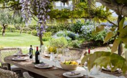 Outdoor dining setup featuring a rustic table adorned with plates, wine glasses, and a bowl of fresh lemons, surrounded by lush greenery and blooming flowers. Wisteria vines hang overhead, creating a serene and inviting atmosphere for al fresco meals.