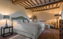 Cozy bedroom featuring a large bed with plush pillows, wooden beams on the ceiling, stylish lamps, and a comfortable seating area, creating a warm and inviting atmosphere.