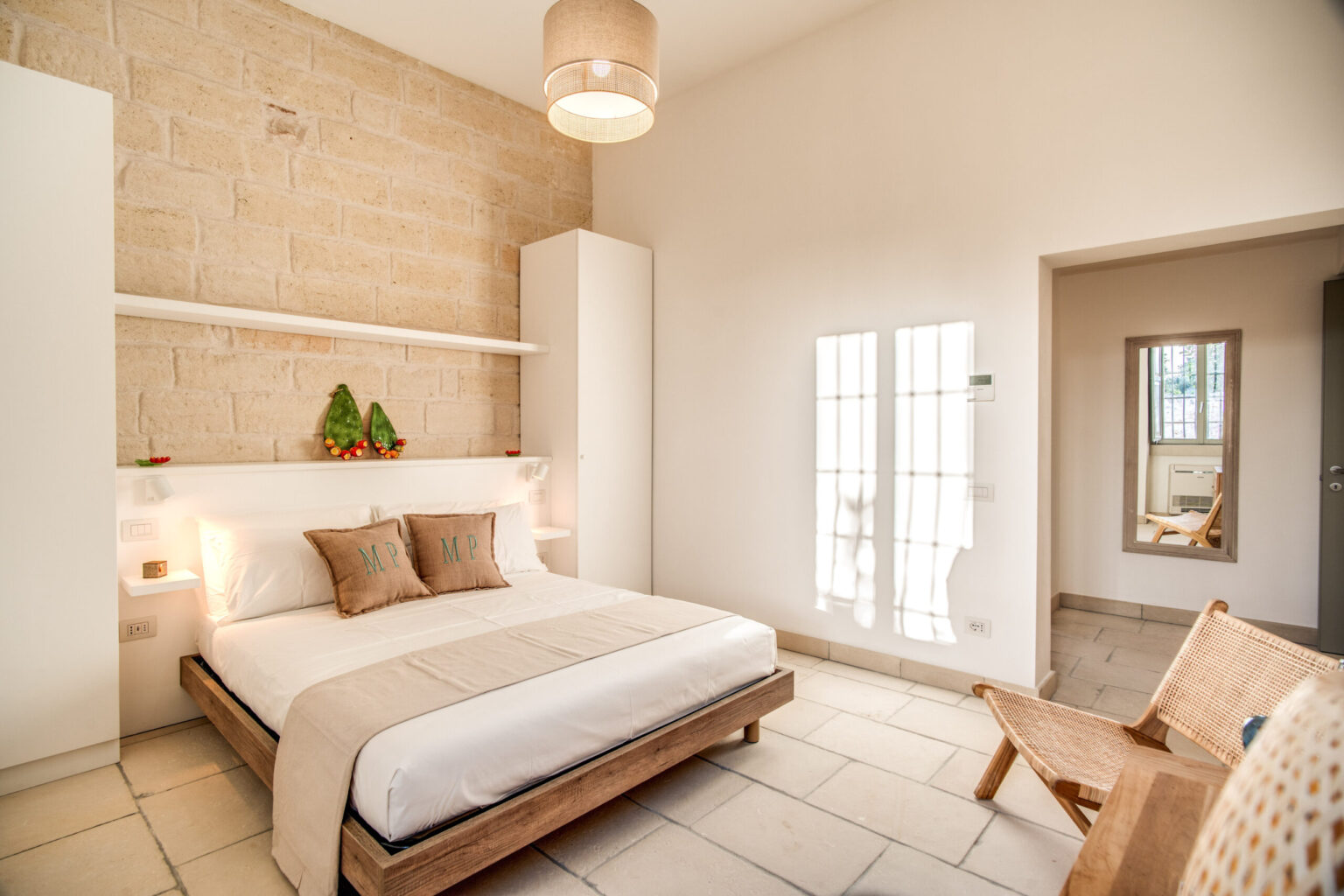 Modern bedroom interior featuring a queen-sized bed with neutral bedding, minimalist decor, natural stone walls, and large windows allowing ample natural light. A wooden chair complements the space, creating a serene and inviting atmosphere. | Only on SopranoVillas