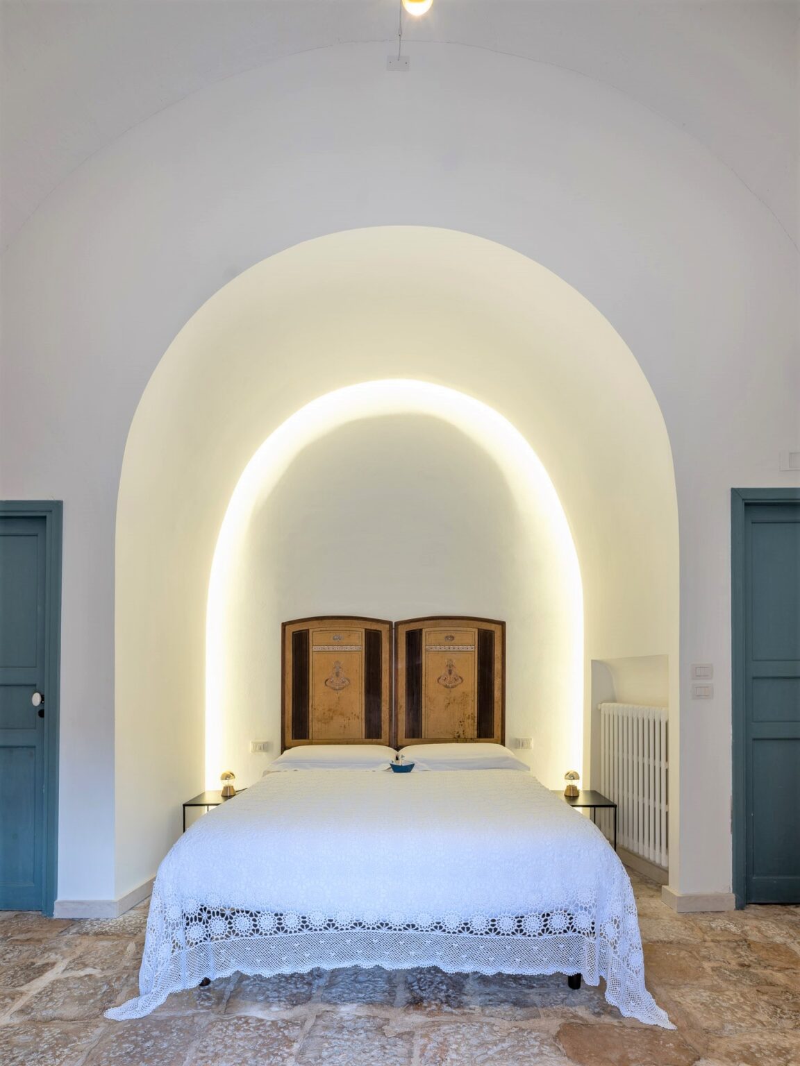 Cozy bedroom featuring a double bed with a white lace bedspread, wooden headboard, and soft ambient lighting. The room has an arched ceiling, stone flooring, and a minimalist decor style, complemented by a blue door and a small nightstand. Ideal for a relaxing getaway. | Only on SopranoVillas