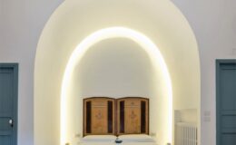 Cozy bedroom featuring a double bed with a white lace bedspread, wooden headboard, and soft ambient lighting. The room has an arched ceiling, stone flooring, and a minimalist decor style, complemented by a blue door and a small nightstand. Ideal for a relaxing getaway.