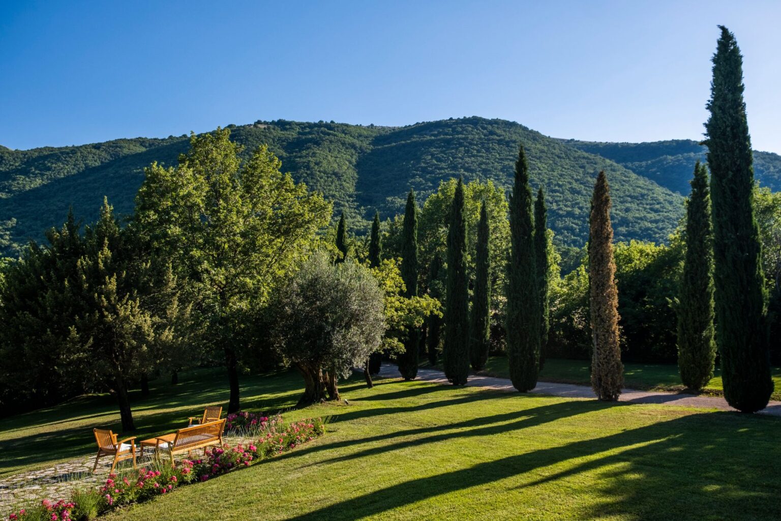 Scenic view of a lush garden with cypress trees and vibrant flowers, set against a backdrop of rolling green hills under a clear blue sky. The image features wooden benches inviting relaxation in a tranquil outdoor setting. | Only on SopranoVillas