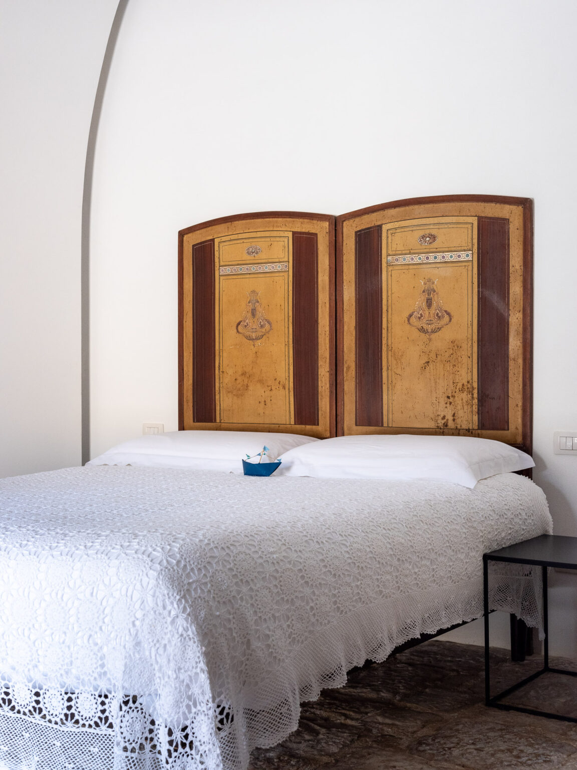 Cozy bedroom featuring a beautifully crafted wooden headboard with intricate designs, a white lace bedspread, and a small decorative boat on the bed. The room has a minimalist aesthetic with neutral walls and a modern black nightstand, creating a tranquil atmosphere. Ideal for relaxation and comfort. | Only on SopranoVillas