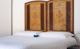 Cozy bedroom featuring a beautifully crafted wooden headboard with intricate designs, a white lace bedspread, and a small decorative boat on the bed. The room has a minimalist aesthetic with neutral walls and a modern black nightstand, creating a tranquil atmosphere. Ideal for relaxation and comfort.