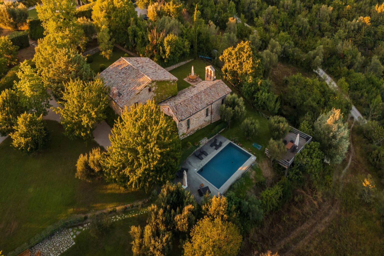 Aerial view of a rustic stone villa surrounded by lush greenery and trees, featuring a swimming pool and outdoor seating area, set in a tranquil countryside landscape. | Only on SopranoVillas