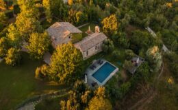 Aerial view of a rustic stone villa surrounded by lush greenery and trees, featuring a swimming pool and outdoor seating area, set in a tranquil countryside landscape.