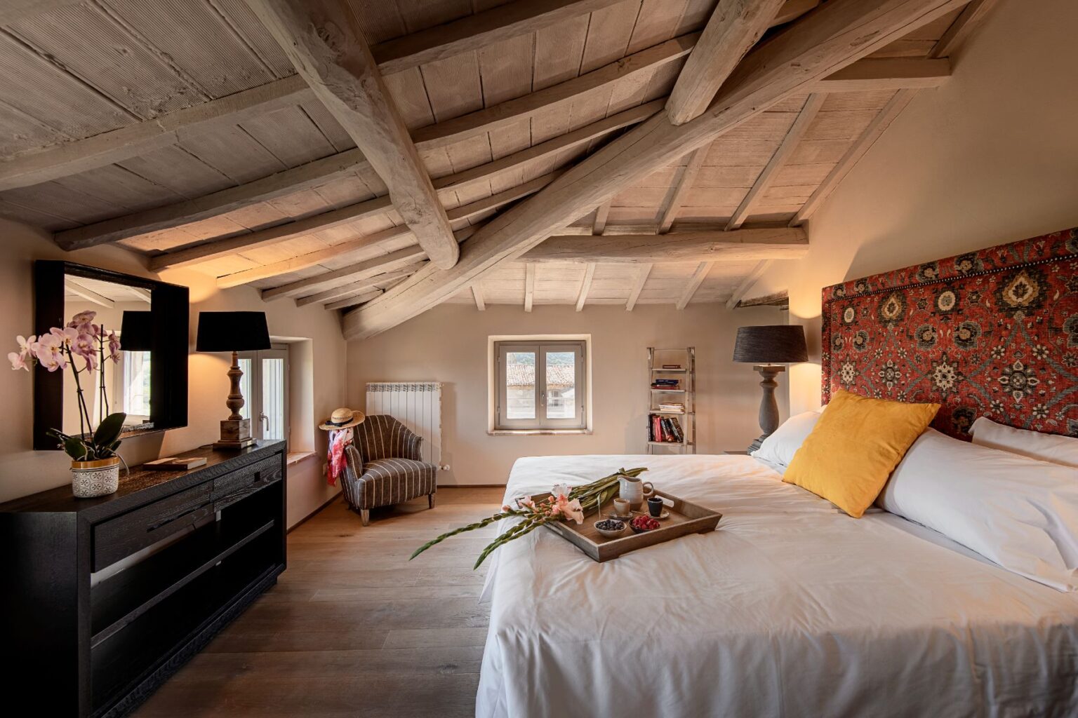Cozy bedroom with sloped wooden ceiling, featuring a large bed with a decorative headboard, a bedside lamp, and a stylish side table. A tray with fresh flowers and snacks sits on the bed. The room includes a vintage armchair, a decorative mirror, and a bookshelf, creating a warm and inviting atmosphere. | Only on SopranoVillas