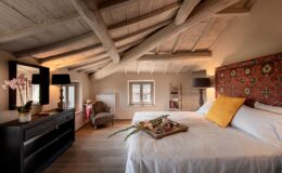 Cozy bedroom with sloped wooden ceiling, featuring a large bed with a decorative headboard, a bedside lamp, and a stylish side table. A tray with fresh flowers and snacks sits on the bed. The room includes a vintage armchair, a decorative mirror, and a bookshelf, creating a warm and inviting atmosphere.