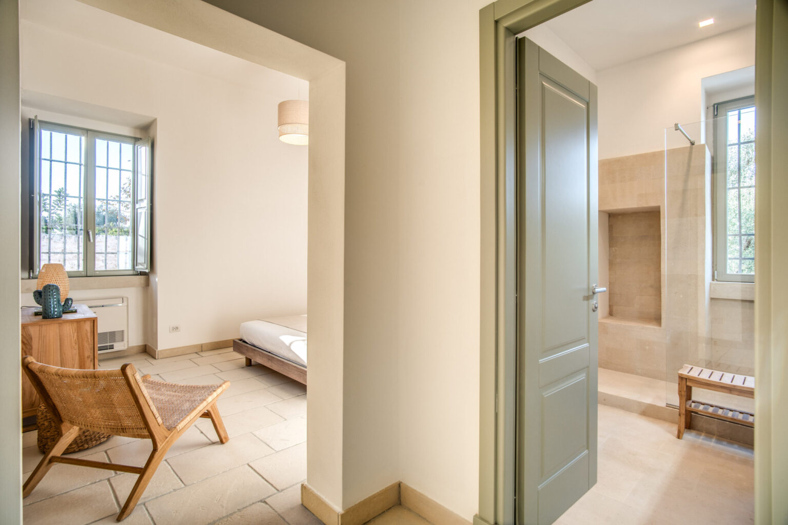 Interior view of a modern bedroom with a wooden chair and a cozy bed, leading to a stylish bathroom featuring a walk-in shower and natural light from windows. | Only on SopranoVillas