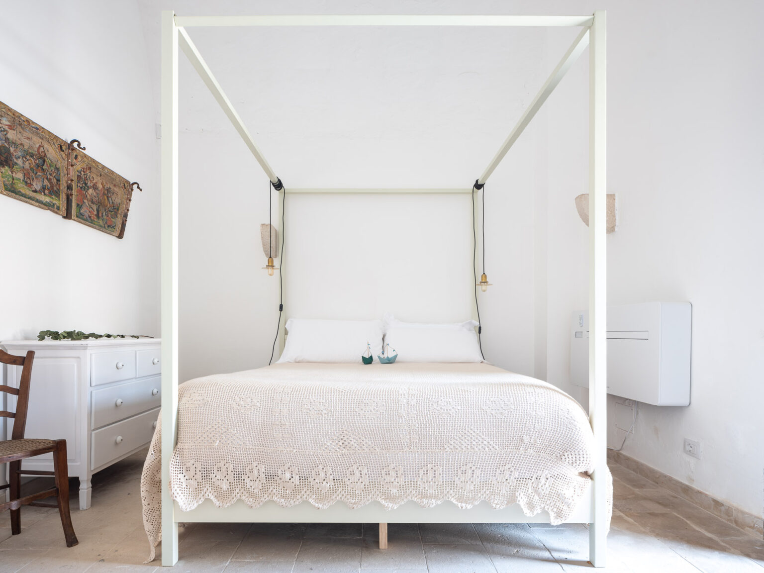 Cozy bedroom featuring a modern four-poster bed with a light-colored crochet blanket, complemented by elegant wall decor and a wooden dresser. The bright, minimalist design creates a serene atmosphere, perfect for relaxation. | Only on SopranoVillas