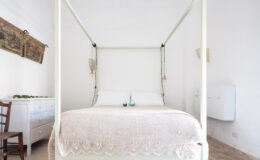 Cozy bedroom featuring a modern four-poster bed with a light-colored crochet blanket, complemented by elegant wall decor and a wooden dresser. The bright, minimalist design creates a serene atmosphere, perfect for relaxation.