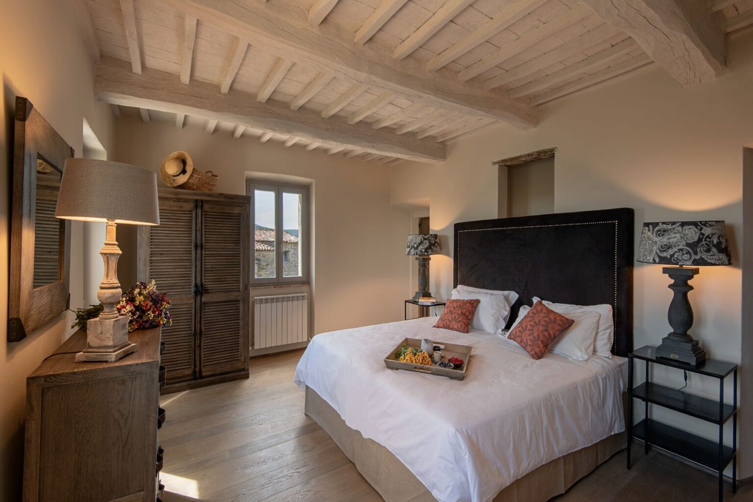 Cozy bedroom featuring a plush king-size bed with decorative pillows, wooden furniture, and stylish lamps. The room boasts a rustic charm with exposed wooden beams, a vintage wardrobe, and a window offering scenic views. A breakfast tray with fresh fruit adds a welcoming touch. Ideal for relaxation and comfort. | Only on SopranoVillas