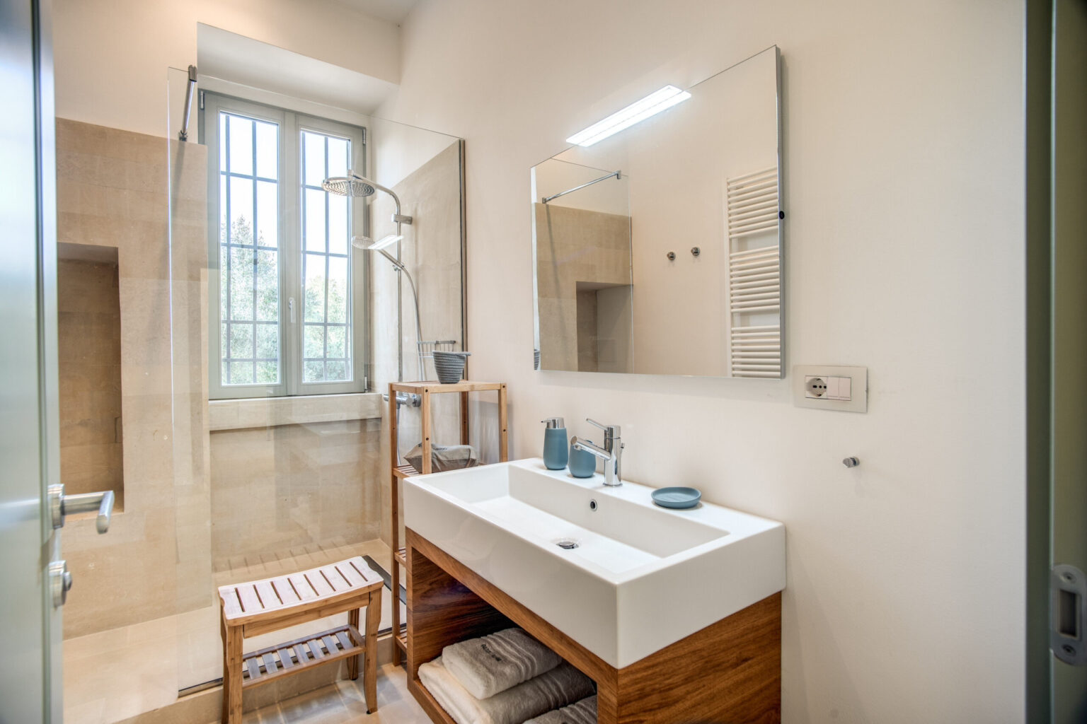 Modern bathroom design featuring a wooden vanity with a white sink, a large mirror, a glass shower enclosure, and natural light from a window. | Only on SopranoVillas
