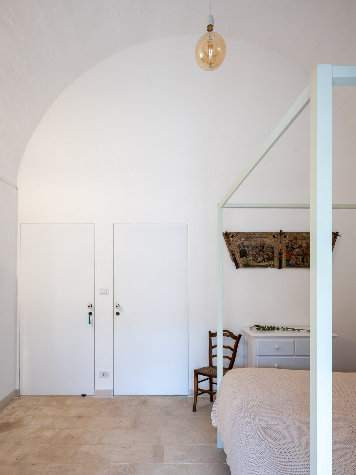 Modern bedroom interior with a high arched ceiling, featuring a four-poster bed, a wooden chair, and a white dresser. The room is well-lit by a vintage-style pendant light, complemented by two closed doors and a decorative wall hanging. | Only on SopranoVillas