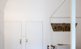 Modern bedroom interior with a high arched ceiling, featuring a four-poster bed, a wooden chair, and a white dresser. The room is well-lit by a vintage-style pendant light, complemented by two closed doors and a decorative wall hanging.