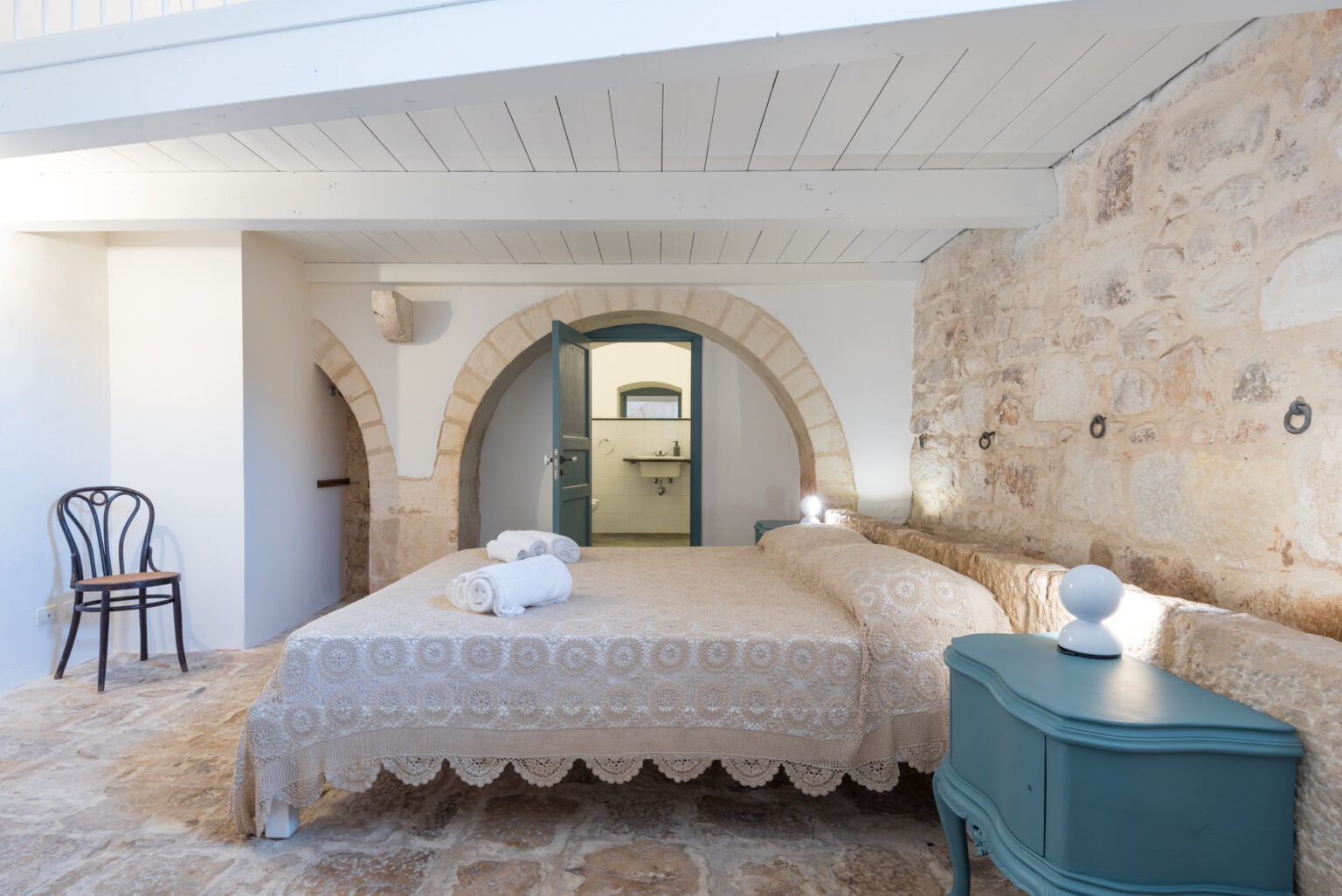 Cozy bedroom featuring a double bed with a decorative beige bedspread, neatly arranged towels, a wooden chair, and a stylish teal nightstand. The room showcases exposed stone walls and wooden beams, with an arched doorway leading to a bathroom. Bright natural light enhances the inviting atmosphere. | Only on SopranoVillas