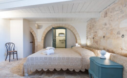 Cozy bedroom featuring a double bed with a decorative beige bedspread, neatly arranged towels, a wooden chair, and a stylish teal nightstand. The room showcases exposed stone walls and wooden beams, with an arched doorway leading to a bathroom. Bright natural light enhances the inviting atmosphere.
