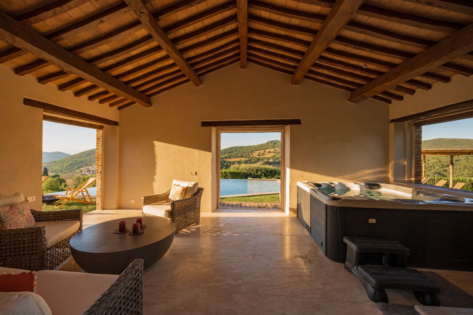 Interior view of a cozy, rustic lounge area featuring comfortable wicker furniture, a round coffee table with candles, and a luxurious hot tub. Large windows showcase stunning views of rolling hills and a serene pool outside, illuminated by warm sunlight. Perfect for relaxation and enjoying nature. | Only on SopranoVillas