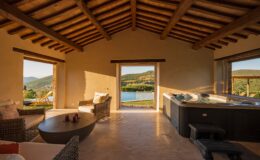 Interior view of a cozy, rustic lounge area featuring comfortable wicker furniture, a round coffee table with candles, and a luxurious hot tub. Large windows showcase stunning views of rolling hills and a serene pool outside, illuminated by warm sunlight. Perfect for relaxation and enjoying nature.