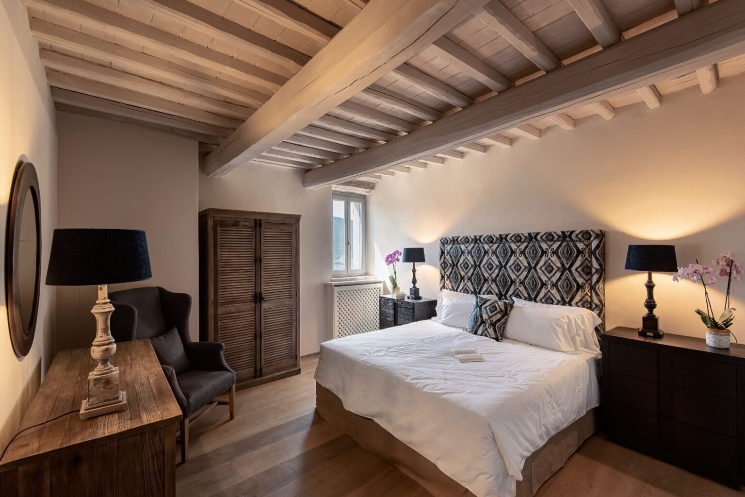 Cozy and modern bedroom featuring a queen-sized bed with a patterned headboard, wooden beams, and stylish decor. The room includes a wooden dresser, a comfortable armchair, and ambient lighting, creating a warm and inviting atmosphere. | Only on SopranoVillas