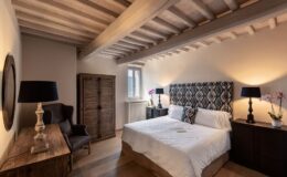 Cozy and modern bedroom featuring a queen-sized bed with a patterned headboard, wooden beams, and stylish decor. The room includes a wooden dresser, a comfortable armchair, and ambient lighting, creating a warm and inviting atmosphere.