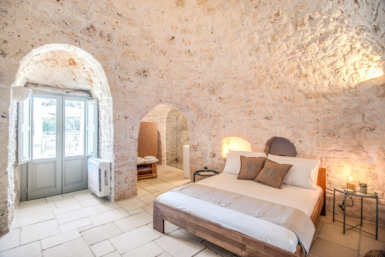 Cozy bedroom with stone walls, featuring a large bed with decorative pillows, side tables, and natural light streaming through arched windows, creating a warm and inviting atmosphere. | Only on SopranoVillas