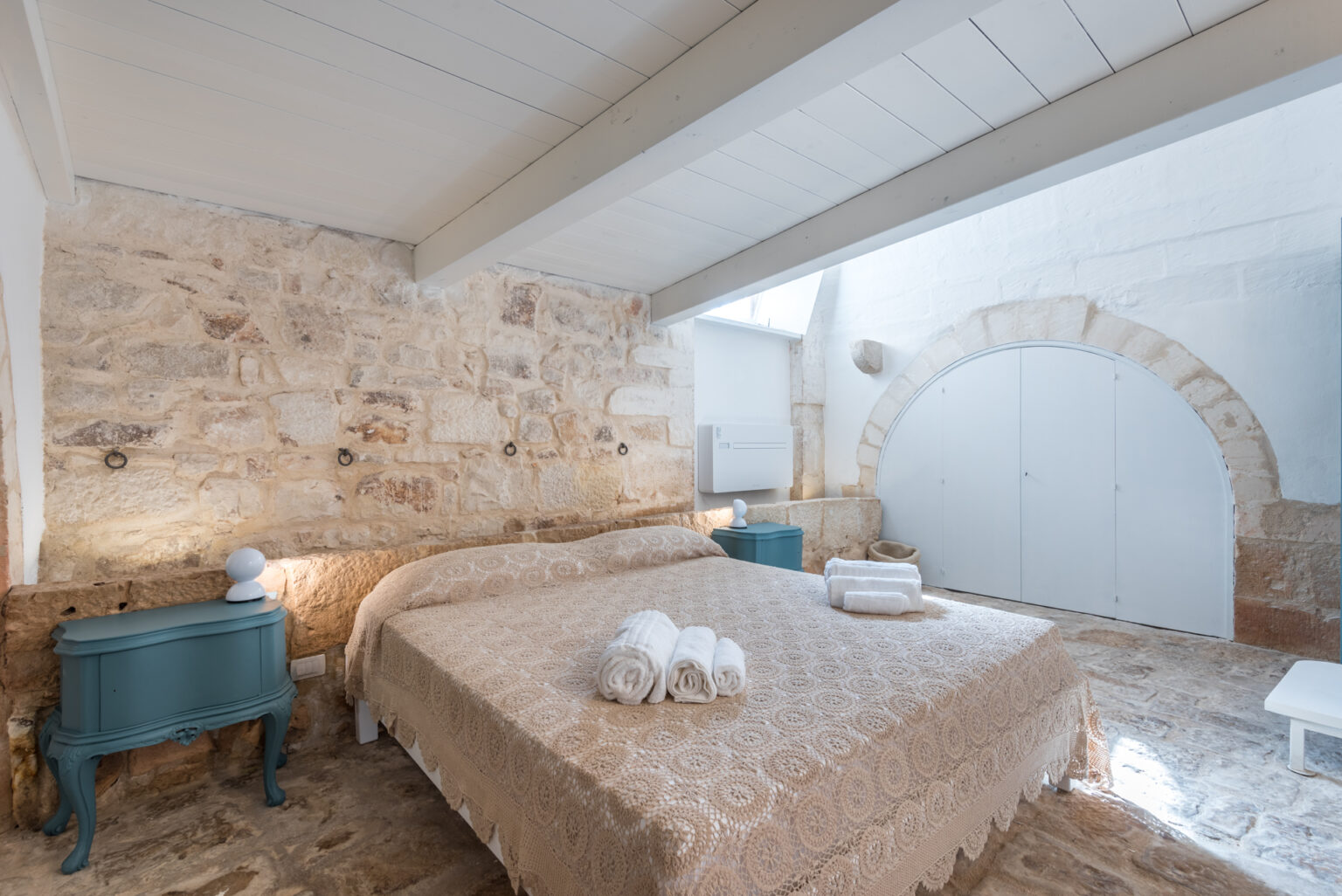 Cozy bedroom featuring a stone wall, a queen-sized bed with a decorative beige coverlet, and neatly arranged white towels. The room includes a vintage blue nightstand with a modern lamp, wooden flooring, and a large arched door in the background, creating a warm and inviting atmosphere. | Only on SopranoVillas