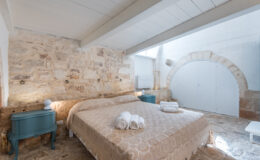 Cozy bedroom featuring a stone wall, a queen-sized bed with a decorative beige coverlet, and neatly arranged white towels. The room includes a vintage blue nightstand with a modern lamp, wooden flooring, and a large arched door in the background, creating a warm and inviting atmosphere.