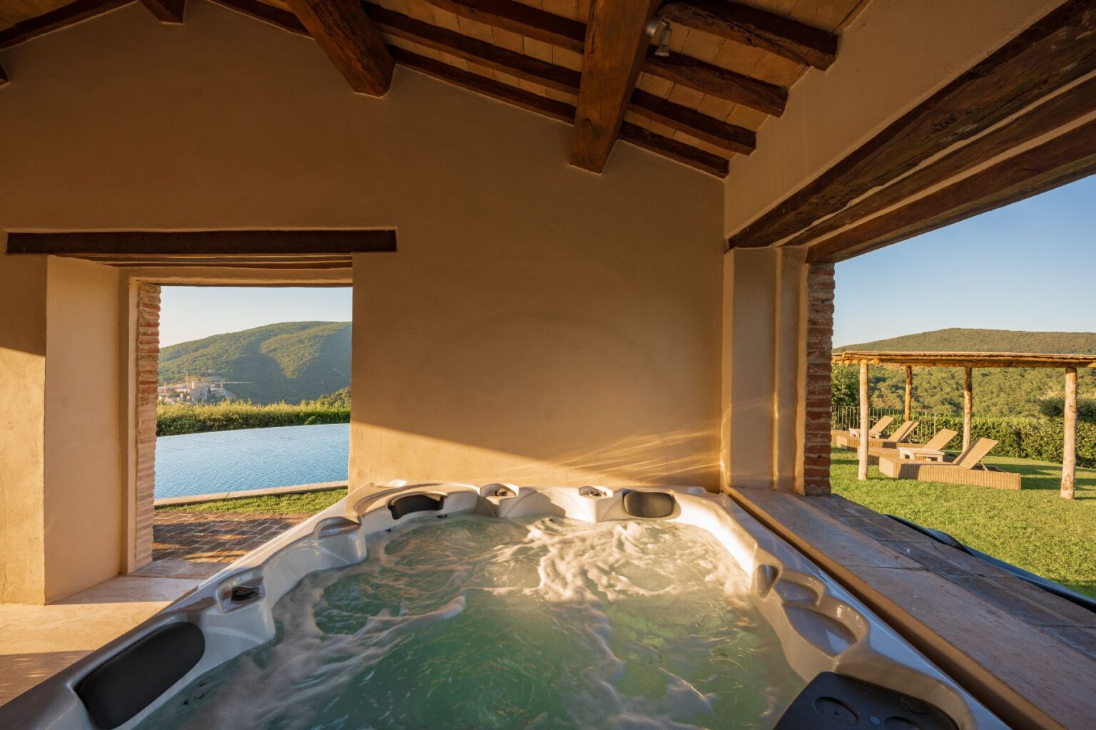 Luxury hot tub with bubbling water inside a rustic villa, featuring large windows that showcase a stunning view of rolling hills and an outdoor pool area with sun loungers. Natural light enhances the serene ambiance. | Only on SopranoVillas