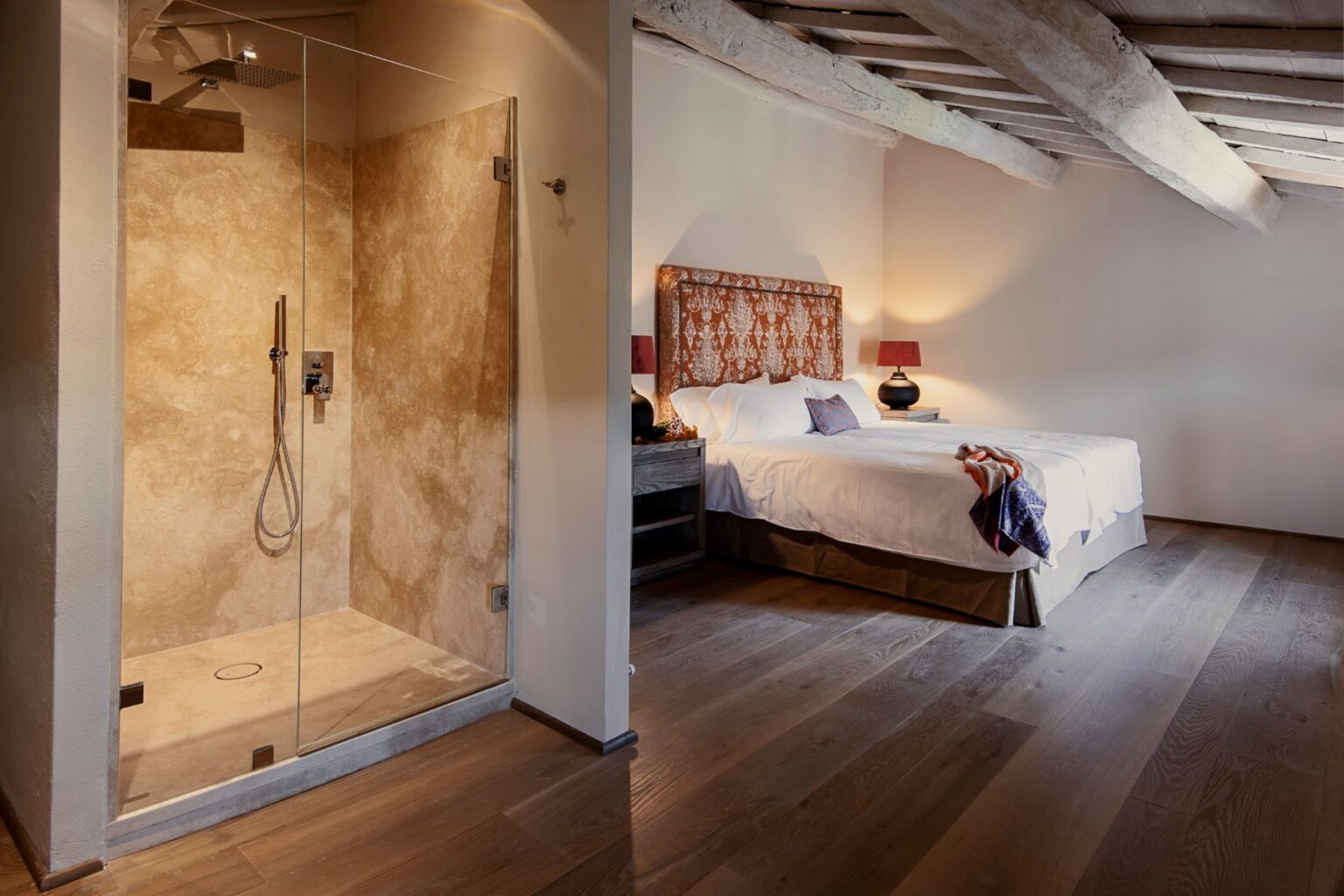 Modern bathroom and bedroom interior featuring a glass shower enclosure with a stone wall, hardwood floors, and a cozy bed with decorative pillows and lamps. Ideal for showcasing stylish home design and relaxation spaces. | Only on SopranoVillas