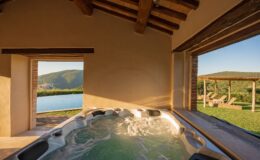 Luxury hot tub with bubbling water inside a rustic villa, featuring large windows that showcase a stunning view of rolling hills and an outdoor pool area with sun loungers. Natural light enhances the serene ambiance.
