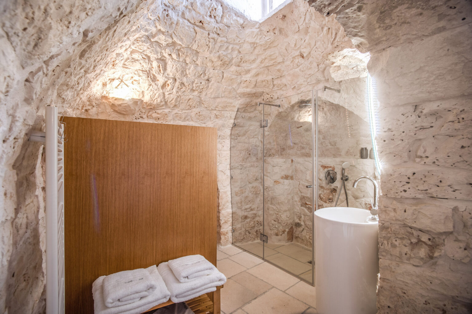Modern bathroom design featuring natural stone walls, a glass shower, and a sleek white pedestal sink, with neatly arranged towels on a wooden bench. | Only on SopranoVillas