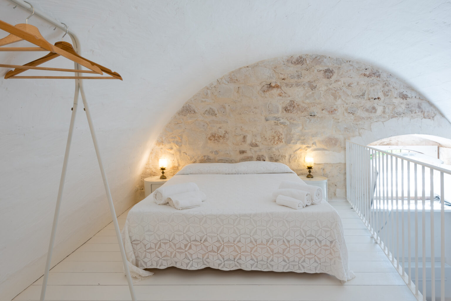 Cozy attic bedroom featuring a double bed with a white patterned blanket, neatly arranged towels, and warm lighting from bedside lamps. The room has a rustic stone wall, wooden hangers on a stylish rack, and a minimalist white railing. Ideal for a relaxing getaway. | Only on SopranoVillas