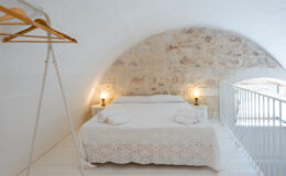 Cozy attic bedroom featuring a double bed with a white patterned blanket, neatly arranged towels, and warm lighting from bedside lamps. The room has a rustic stone wall, wooden hangers on a stylish rack, and a minimalist white railing. Ideal for a relaxing getaway.