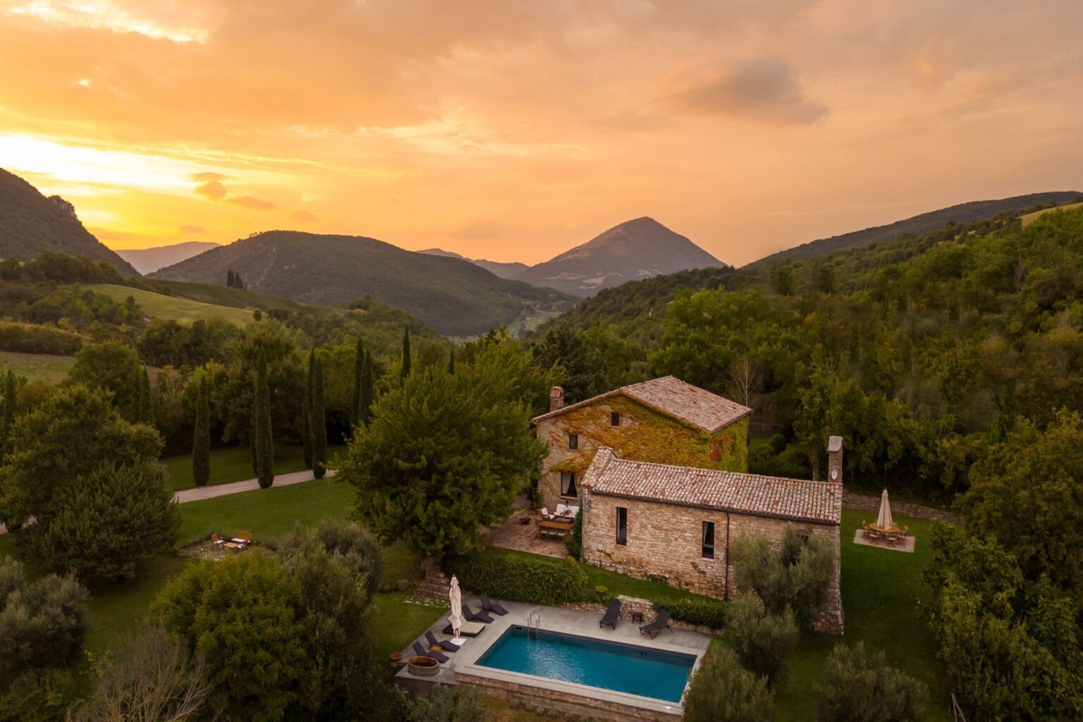 Scenic view of a rustic stone villa with a swimming pool, surrounded by lush greenery and rolling hills at sunset, showcasing the tranquility of countryside living. | Only on SopranoVillas
