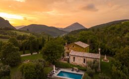 Scenic view of a rustic stone villa with a swimming pool, surrounded by lush greenery and rolling hills at sunset, showcasing the tranquility of countryside living.