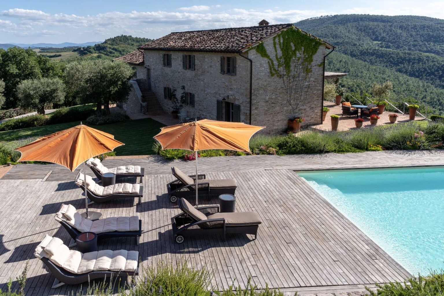 Luxurious villa in a scenic landscape with a swimming pool, wooden deck, and sun loungers under orange umbrellas, surrounded by lush greenery and rolling hills. | Only on SopranoVillas