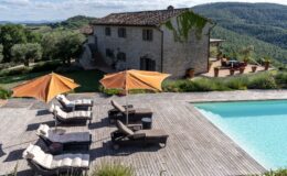 Luxurious villa in a scenic landscape with a swimming pool, wooden deck, and sun loungers under orange umbrellas, surrounded by lush greenery and rolling hills.